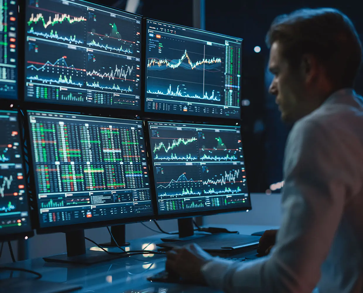Person analyzing multiple financial charts and graphs on computer screens in a dimly lit office.