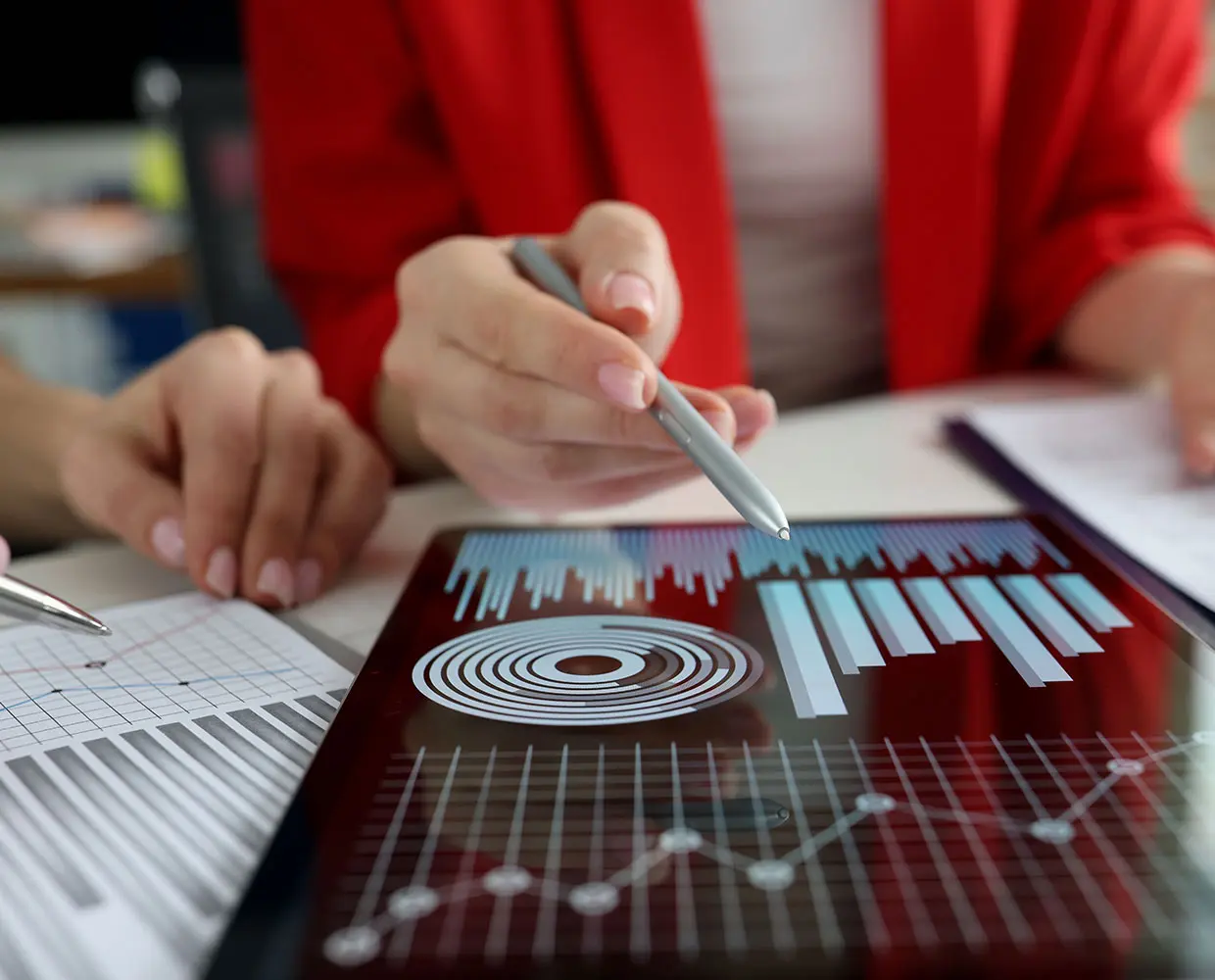 Two people discussing colorful bar graphs and a pie chart on a tablet screen, with papers showing data charts on the table.