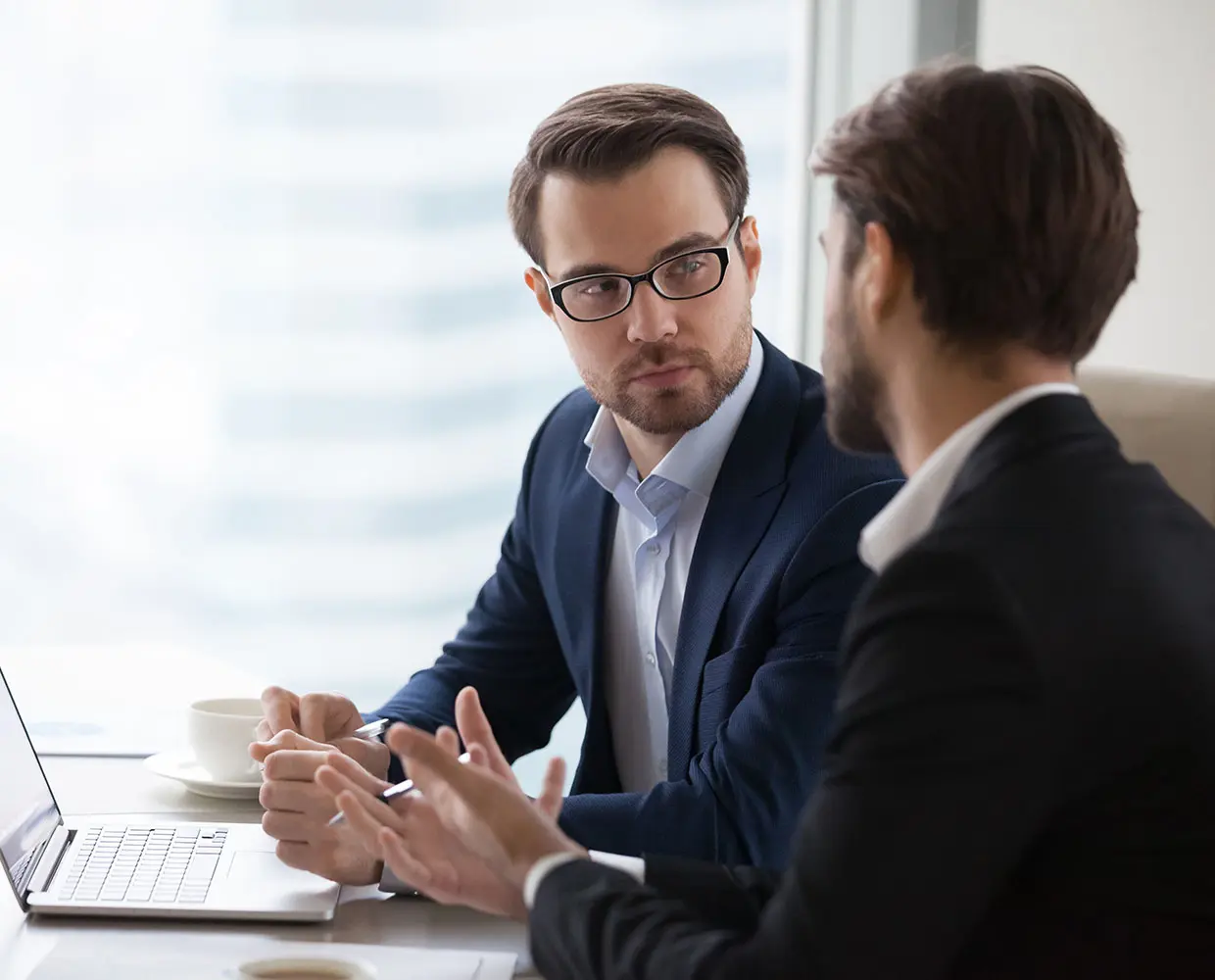 Two men in business attire having a serious discussion at a desk with a laptop and coffee cups.