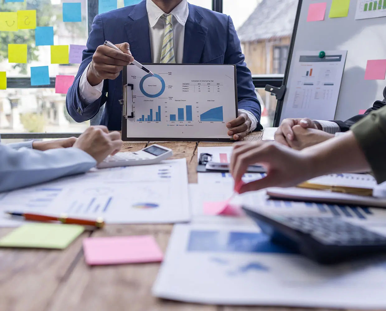 Business meeting with people discussing charts and graphs, one person pointing to a clipboard with a pie chart. Various documents and a calculator are on the table.