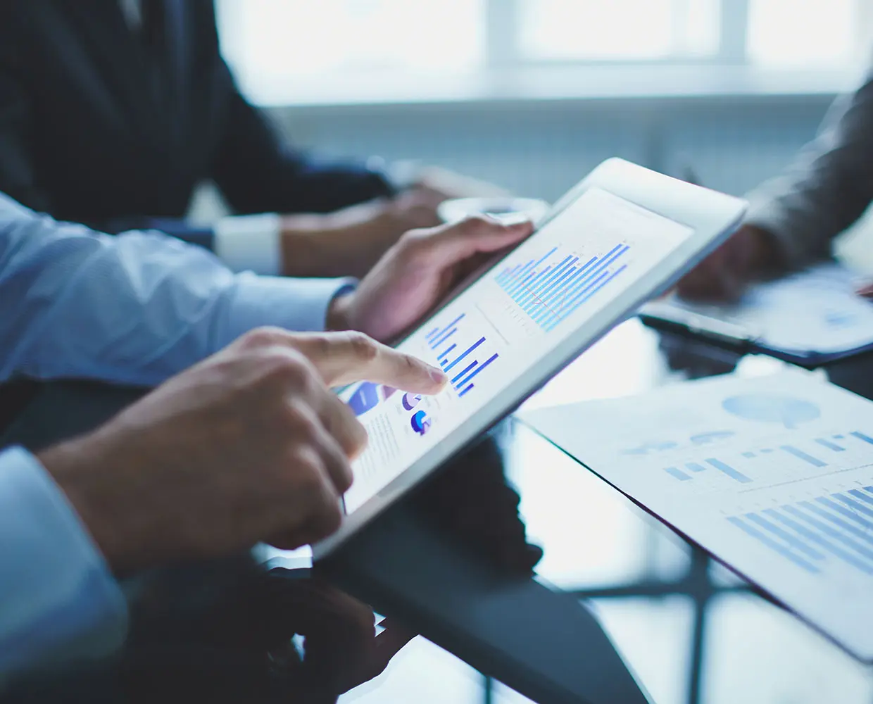 Person in a suit pointing at a bar chart on a tablet in an office setting, with printed graphs on the table.