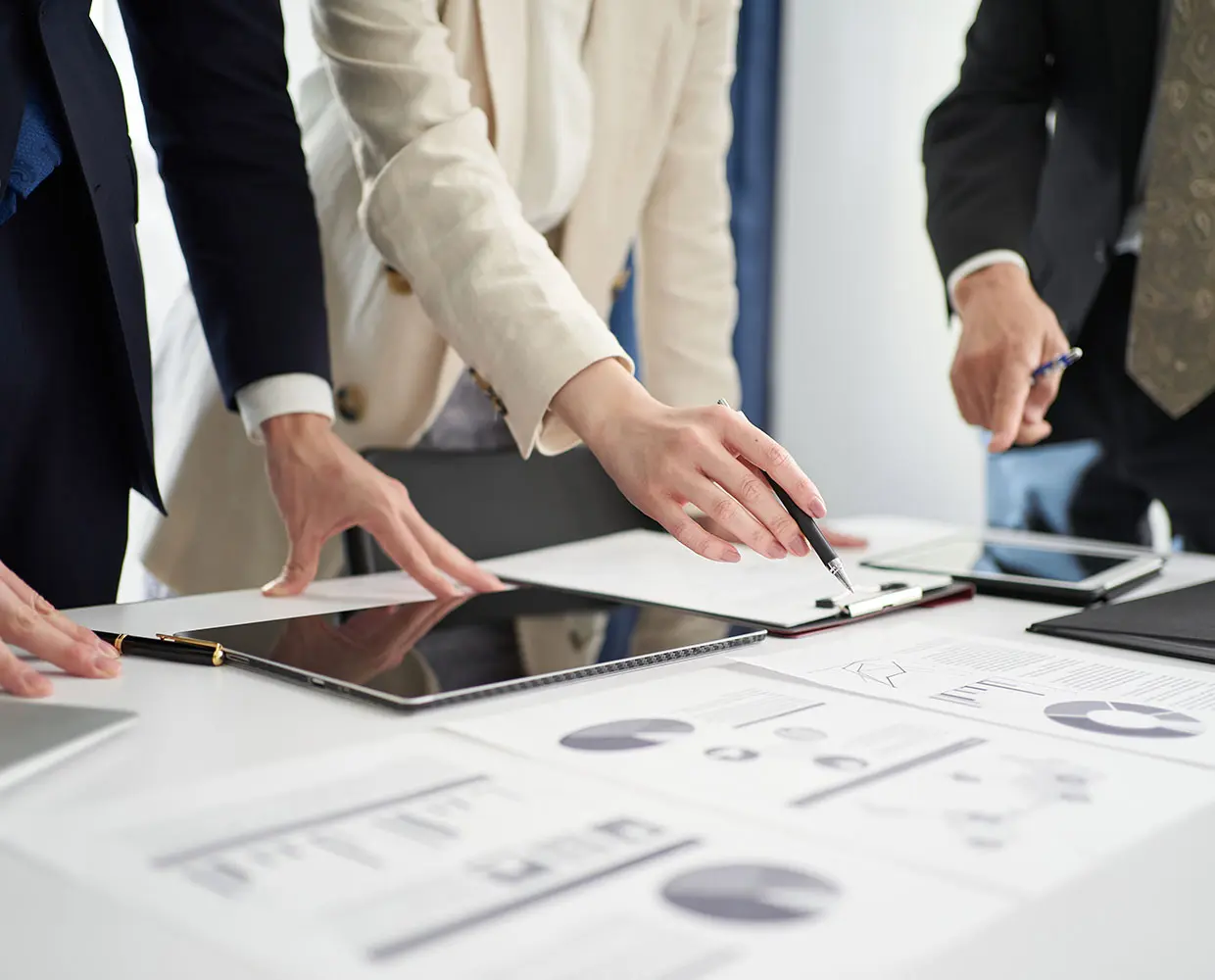 Three people in business attire review documents and charts on a table, with one pointing to a data sheet using a pen.