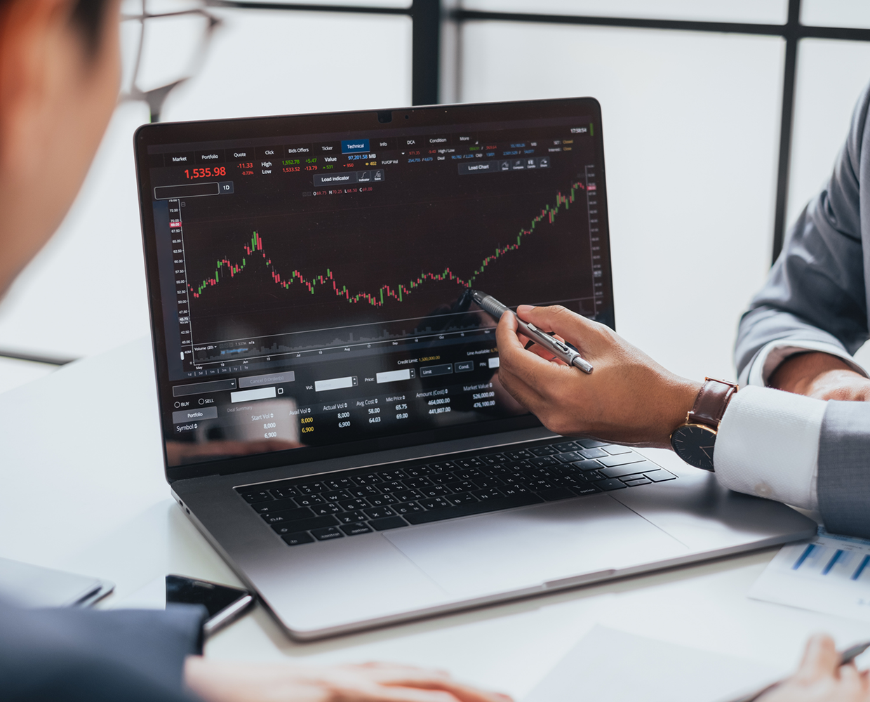 Two individuals discuss a stock market graph on a laptop screen, with one person pointing at the chart using a pen.