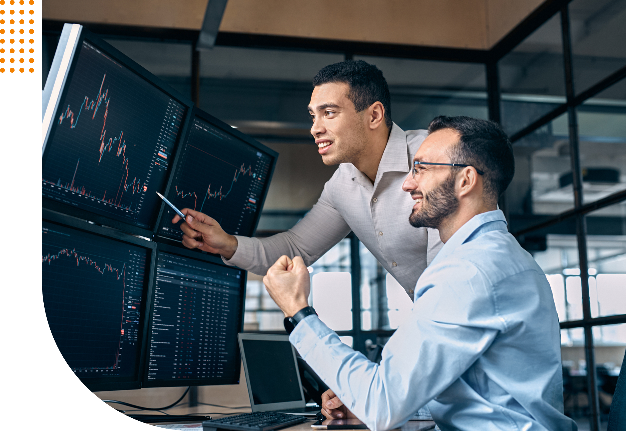 Two men look at financial charts on multiple monitors in an office. One points at the screens while the other holds a fist up in excitement.