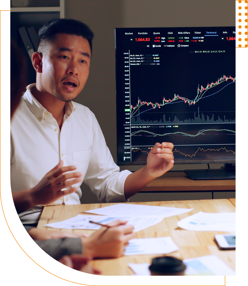 Man in white shirt gestures while discussing financial charts displayed on a screen during a meeting.