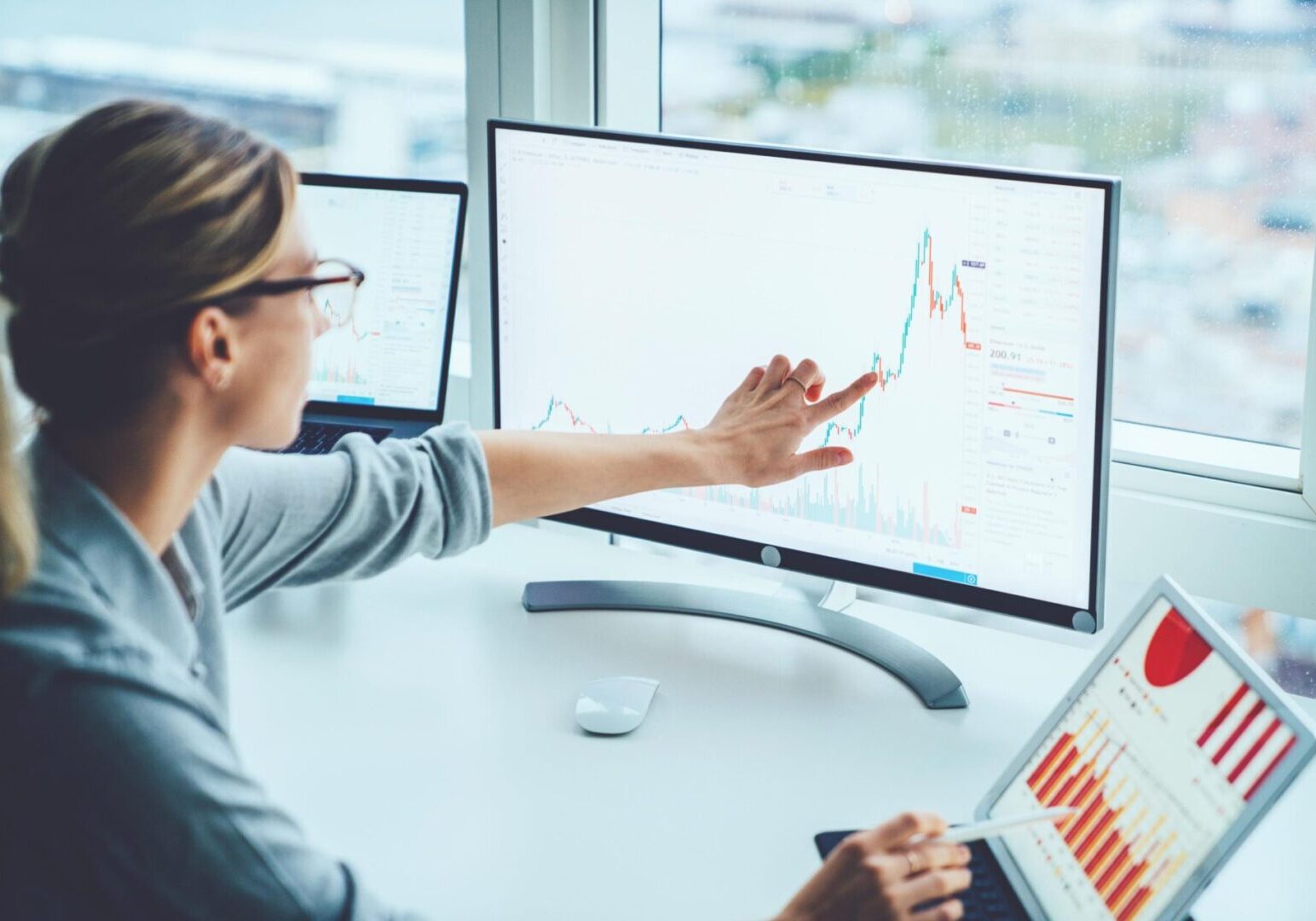 A woman in glasses points at a financial chart on a computer monitor, with graphs also displayed on a laptop and an additional screen.