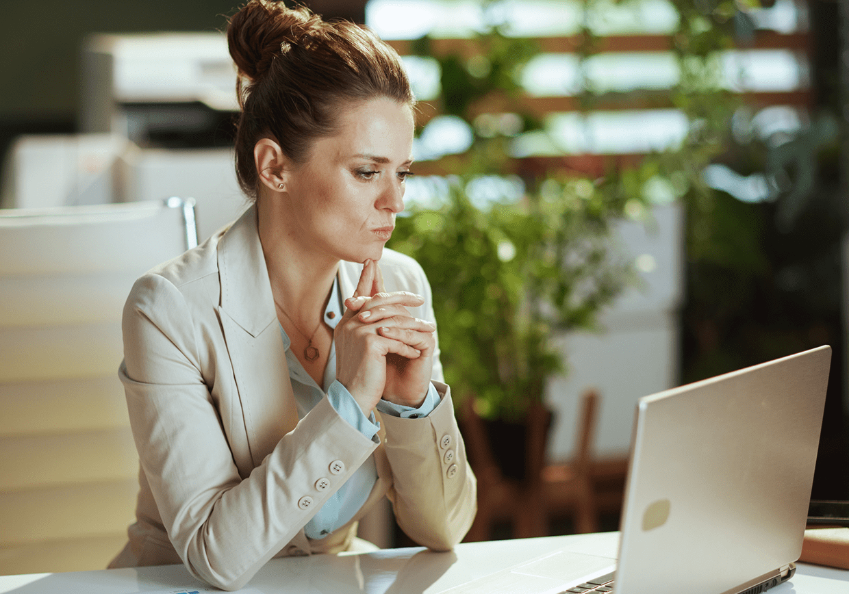 A woman in a blazer sits at a desk, looking at a laptop screen with a thoughtful expression, hands clasped under her chin. Papers with graphs are on the desk. Plants are in the background.