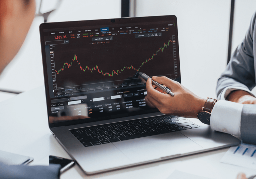 Two individuals discuss a stock market graph on a laptop screen, with one person pointing at the chart using a pen.